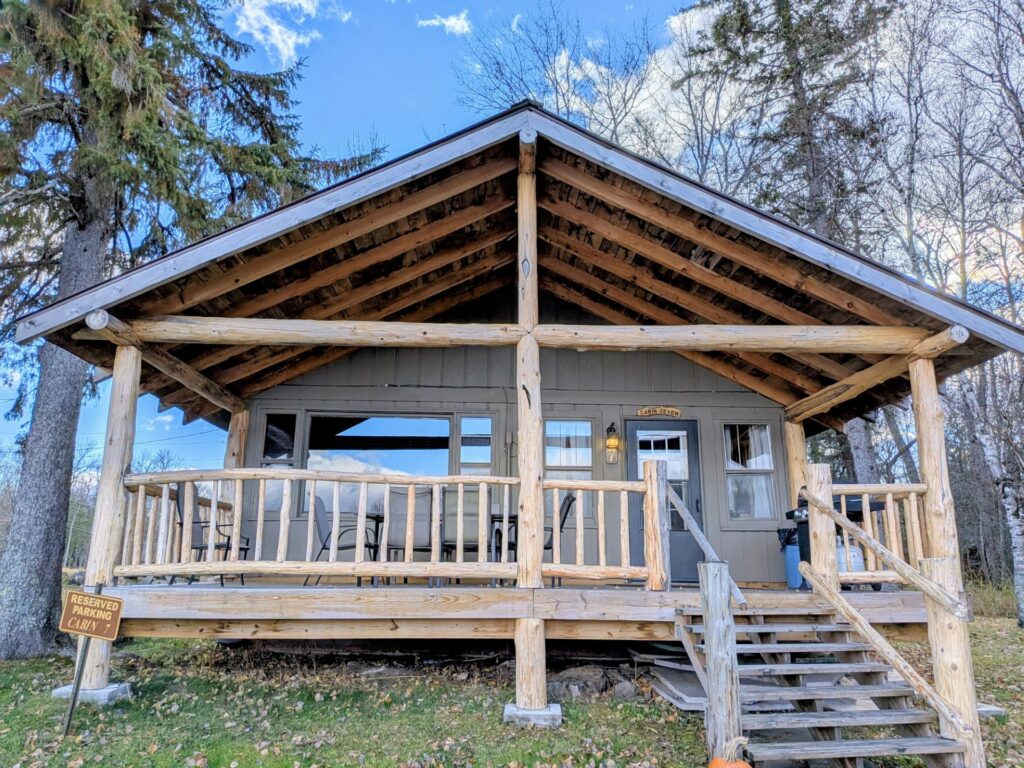 A rustic wooden cabin with a covered porch, railings, and steps sits among trees, evoking the spirit of Thunderbird country. A “Reserved Parking” sign is visible in front, and the sky above is mostly clear with a few clouds. | Rainy Lake, MN