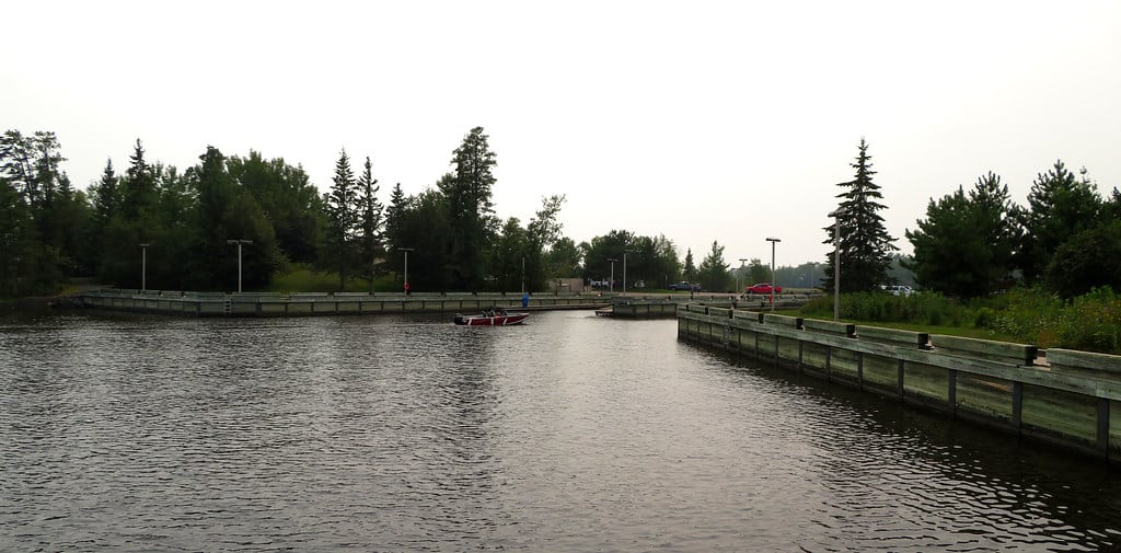 A small dock area with wooden railings surrounds calm water on Rainy Lake, bordered by green trees and shrubs. A red boat is moored at the dock, and a few cars are visible in the distance under Minnesota’s overcast sky. | Rainy Lake, MN