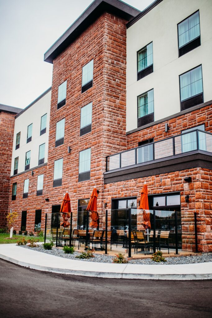 A modern hotel in Minnesota with red brick and white walls, featuring large windows and an outdoor patio area with tables, chairs, and closed orange umbrellas—perfect after exploring nearby Rainy Lake. | Rainy Lake, MN