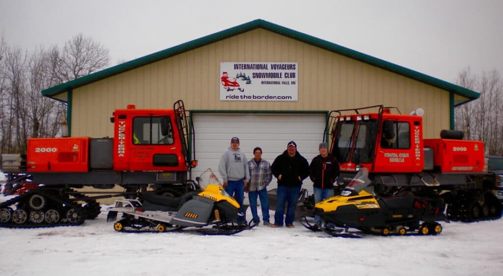 Four people stand in front of two large red snow groomers and two snowmobiles outside a building with a sign reading "International Voyageurs Snowmobile Club" on a snowy day near Rainy Lake, Minnesota. | Rainy Lake, MN