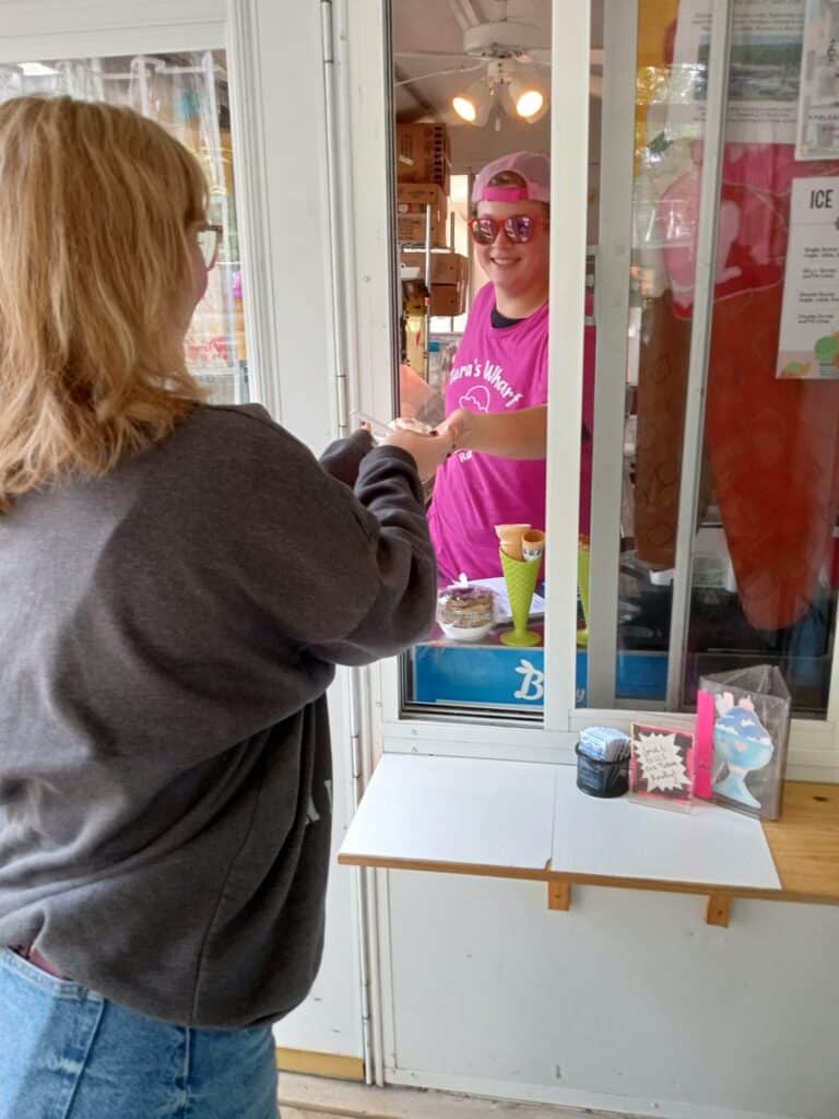 A person in a gray sweatshirt receives ice cream from a smiling worker in a pink shirt and cap at a Rainy Lake walk-up window. The worker is handing over a cone through the window counter, bringing sweet joy to this Minnesota moment. | Rainy Lake, MN