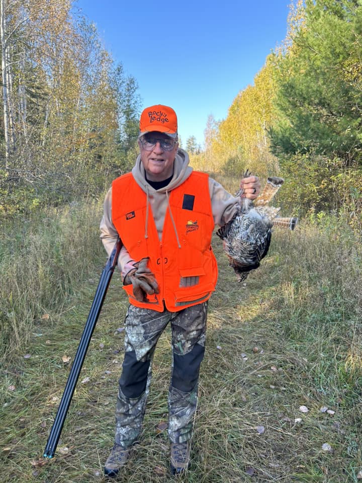 A person wearing camouflage pants, a tan hoodie, and an orange hunting vest and cap stands on a grassy path near Minnesota’s Rainy Lake, holding a shotgun and a wild turkey, surrounded by trees under a clear blue sky. | Rainy Lake, MN