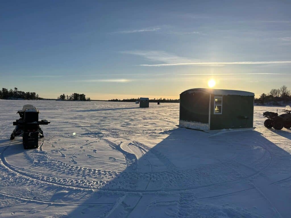 A snow-covered frozen lake at sunrise in Minnesota, with a snowmobile and a green ice fishing hut in the foreground. Two more huts and tire tracks mark the icy surface, with trees lining the distant horizon—classic Rainy Lake winter scenery. | Rainy Lake, MN
