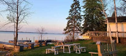 Wooden picnic tables and benches sit on a grassy area by Rainy Lake at sunset, with docks, trees, and a white cabin nearby. The calm Minnesota water reflects the soft evening light.