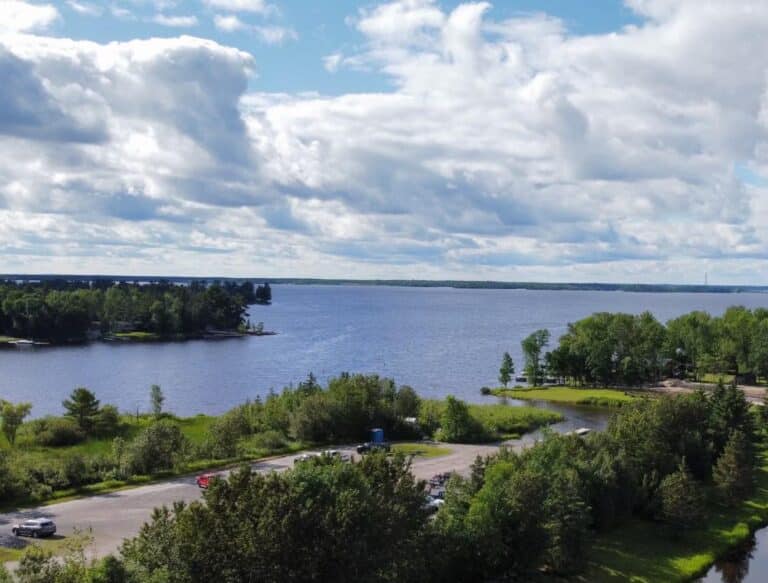 A scenic lakeside view of Rainy Lake in Minnesota, with a partly cloudy sky, trees and greenery along the shore, and a parking lot with several cars in the foreground. | Rainy Lake, MN
