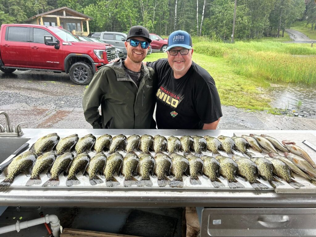 Two men smiling and posing behind a table covered with many freshly caught fish, arranged in neat rows. A red truck and cabin sit nearby, surrounded by green trees—capturing a perfect day of fishing at Rainy Lake, Minnesota. | Rainy Lake, MN