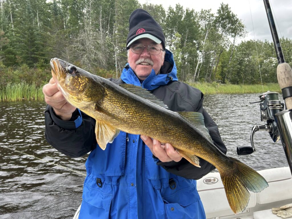 A man in a blue jacket and black hat smiles while holding a large fish on a boat. Trees and water are visible in the background, suggesting a lakeside fishing trip on Rainy Lake in Minnesota. | Rainy Lake, MN