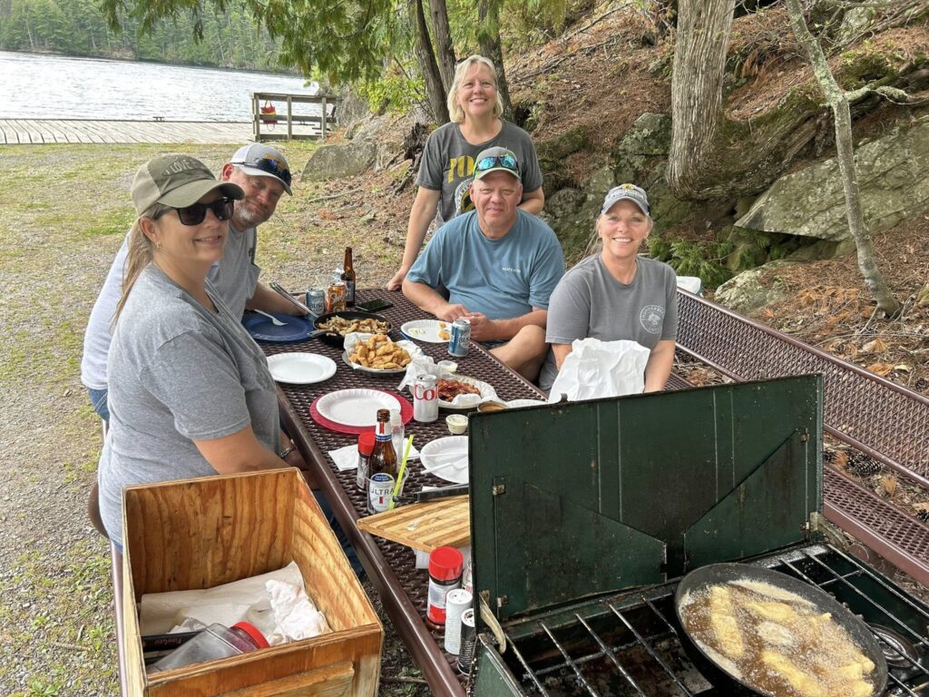 Five adults smile at a picnic table by Rainy Lake, enjoying a meal of fried food. A portable stove sizzles in the foreground as trees and water create a peaceful Minnesota lakeside setting. | Rainy Lake, MN