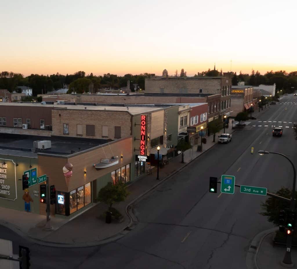 A small town street in Minnesota at sunset with brick buildings, shops, and a neon sign reading “Ronning’s.” The quiet streets glow under green lights, and an ice cream cone sign stands on a corner store near Rainy Lake. | Rainy Lake, MN