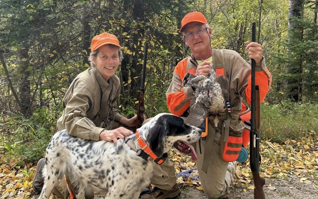 Two people in hunting gear and orange hats pose with a hunting dog in a Minnesota forest. One holds a rifle and game bird, while the other kneels, smiling with a hand on the dog. Fallen leaves cover the ground. | Rainy Lake, MN
