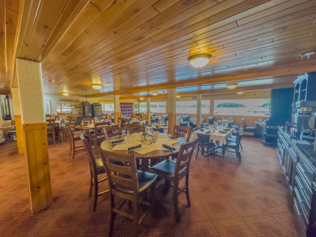 A cozy restaurant interior with wooden ceiling and walls, set tables with chairs, dim lighting, and large windows showing trees outside reflects the welcoming spirit of Thunderbird hospitality. | Rainy Lake, MN