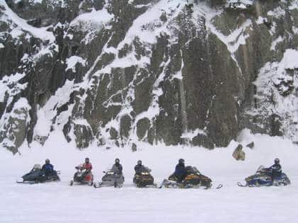 Six people on snowmobiles are lined up on a snowy surface in front of a rocky, snow-covered cliff at Rainy Lake, Minnesota. The riders are dressed in winter gear, highlighting the cold and wintry scene. | Rainy Lake, MN