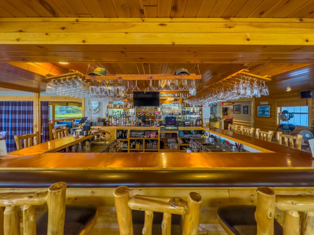 A rustic wooden bar interior with hanging glasses, bottles on shelves, and wooden stools, featuring a central square bar counter and a TV in the background. Warm lighting creates a cozy atmosphere reminiscent of the classic Thunderbird lounge experience. | Rainy Lake, MN