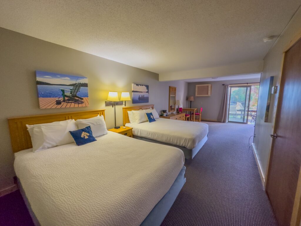 A Thunderbird hotel room with two double beds, a nightstand, wall art, desk, and chairs. There is a sliding glass door leading to a balcony with a view of trees outside. | Rainy Lake, MN