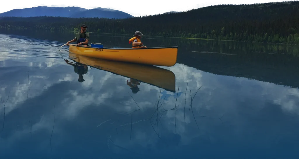 A yellow canoe with an adult and child glides across the calm, blue waters of Rainy Lake, Minnesota. Mountains and trees line the background as the water reflects the sky and clouds, creating a serene scene. | Rainy Lake, MN
