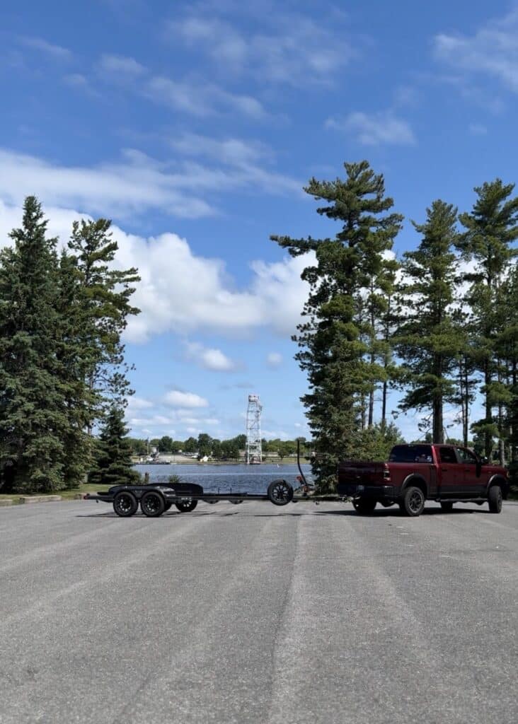 A maroon pickup truck with an empty boat trailer is parked near a lakeside boat launch on Rainy Lake, Minnesota, surrounded by tall pine trees under a blue sky with scattered clouds. | Rainy Lake, MN