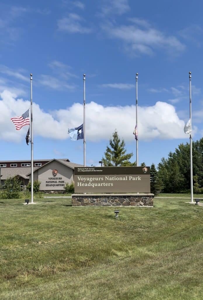 Five flags at half-staff stand behind a sign reading "Voyageurs National Park Headquarters" near Rainy Lake in Minnesota, set before a stone building and green lawn, with trees and a partly cloudy blue sky in the background. | Rainy Lake, MN