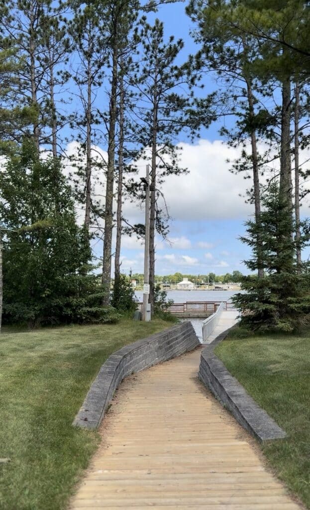 A wooden pathway bordered by low stone walls leads through tall trees and grass toward Rainy Lake in Minnesota, with a dock and buildings visible across the water under a partly cloudy sky. | Rainy Lake, MN