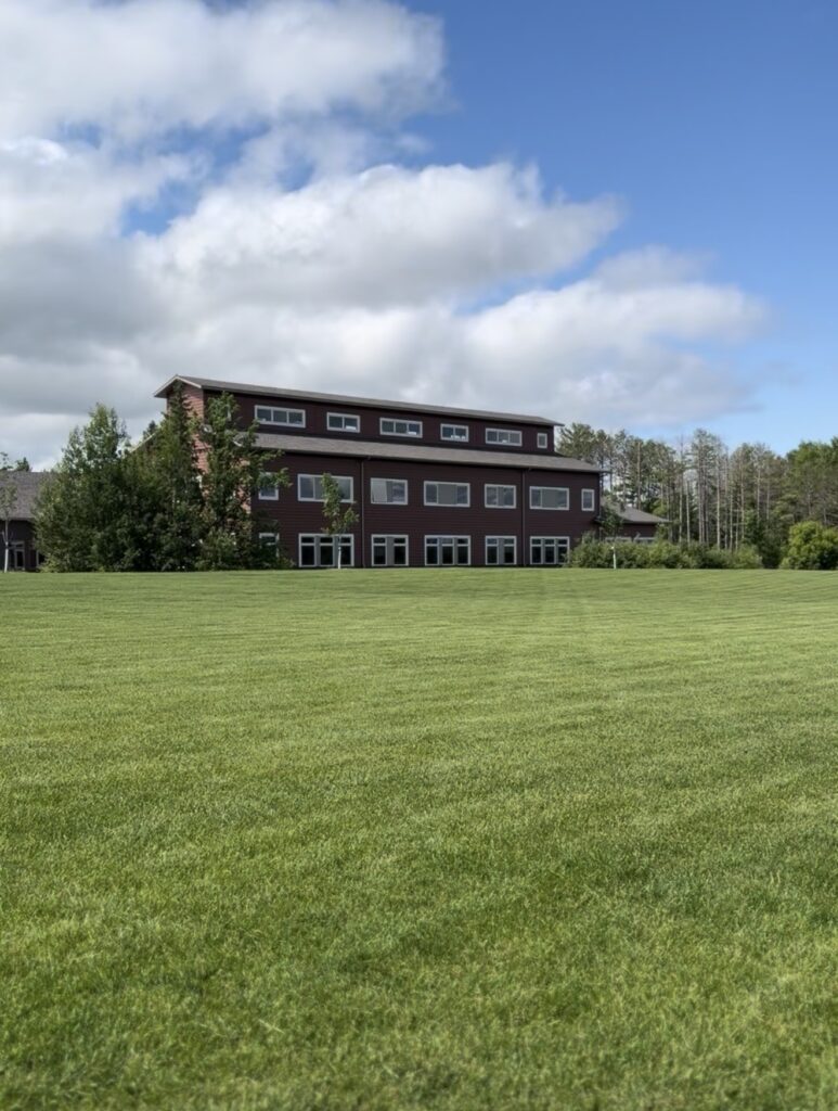 A large brown building with many windows sits behind a wide, neatly mowed green lawn under a partly cloudy blue Minnesota sky, surrounded by trees near Rainy Lake. | Rainy Lake, MN