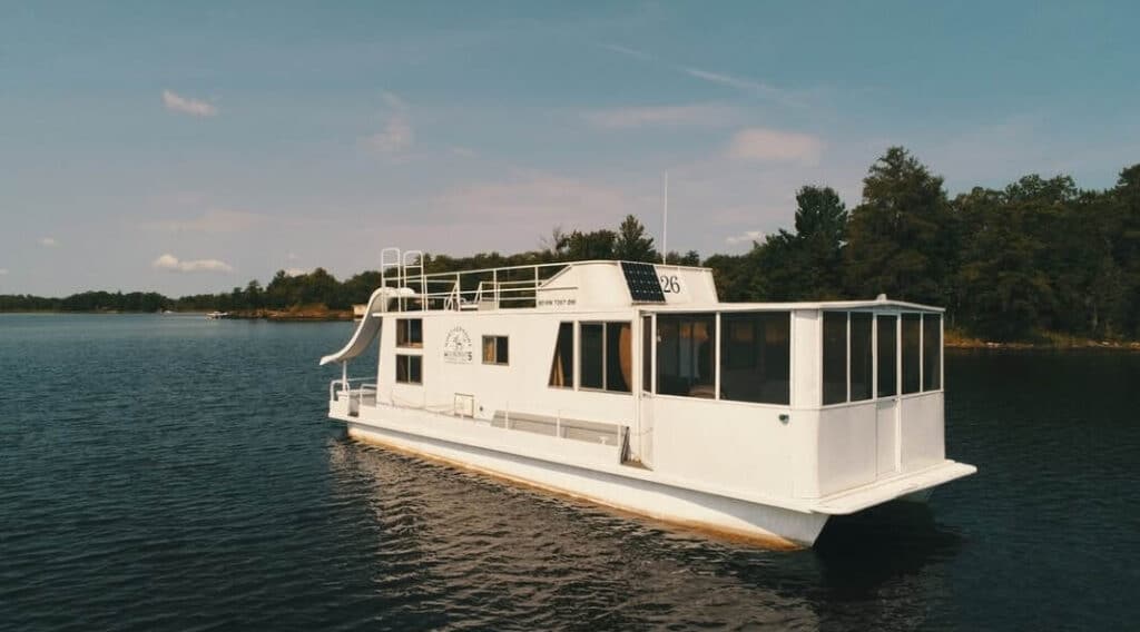 A white houseboat with a waterslide on its roof floats on the calm waters of Rainy Lake in Minnesota, surrounded by trees and a clear sky in the background.