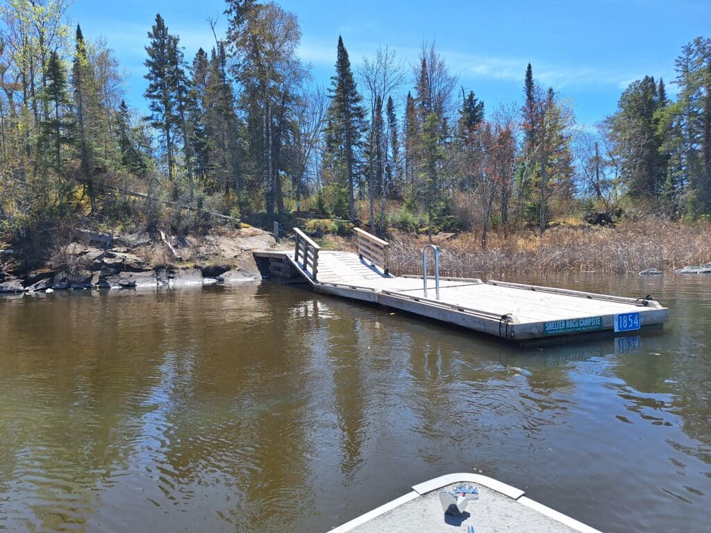 A wooden dock extends over calm water near Shelter Rock, surrounded by trees and brush. The photo is taken from a boat approaching the dock under a bright blue sky, hinting at nearby campsites nestled in the wilderness. | Rainy Lake, MN