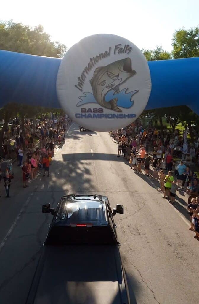 Aerial view of a black truck passing under a blue inflatable arch with an "International Falls Bass Championship" logo, surrounded by large crowds lining the street at this popular Minnesota event near scenic Rainy Lake. | Rainy Lake, MN
