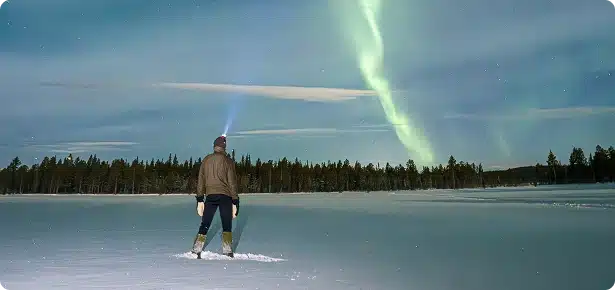 A person wearing boots and a jacket stands on a snowy field at night near Rainy Lake, Minnesota, facing green Northern Lights in the sky. Trees are visible in the distance as a beam of light shines from their headlamp.