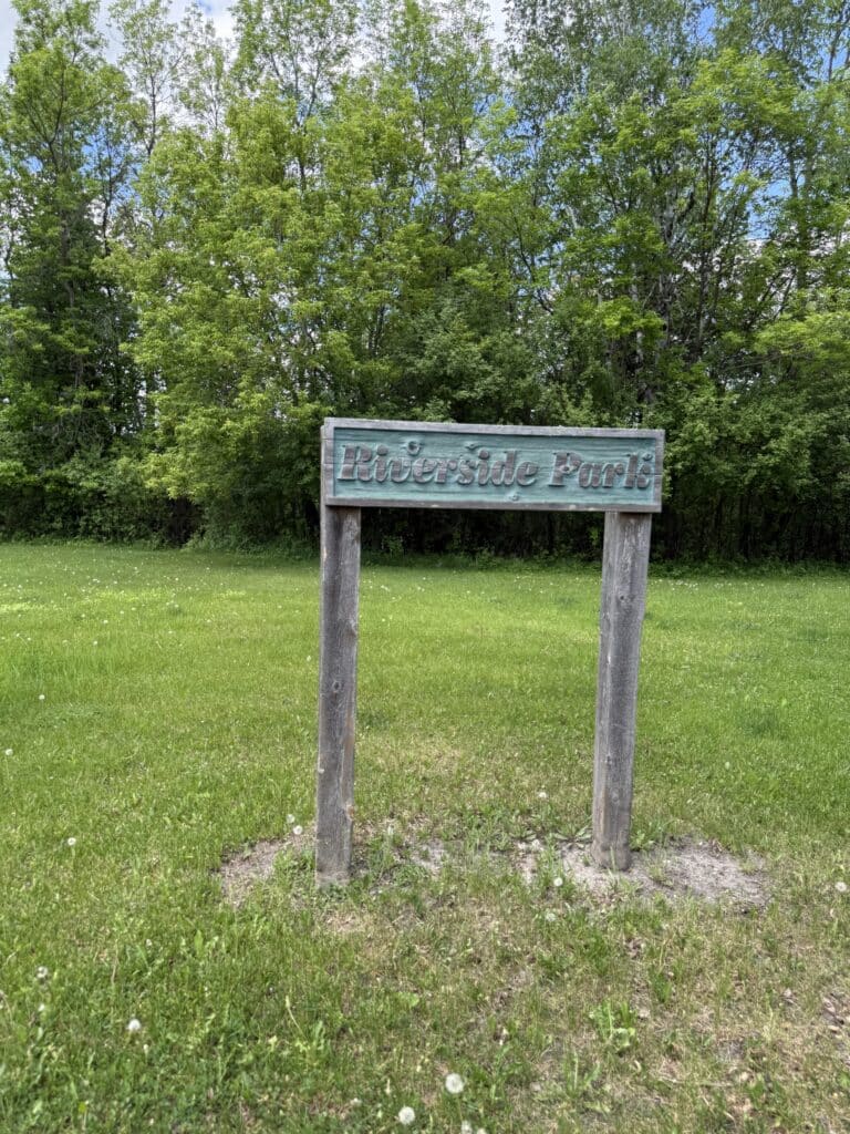 A weathered wooden sign reading "Riverside Park" stands on a grassy field, inviting visitors to explore the lush green trees beneath a cloudy blue sky in Riverside Park. | Rainy Lake, MN