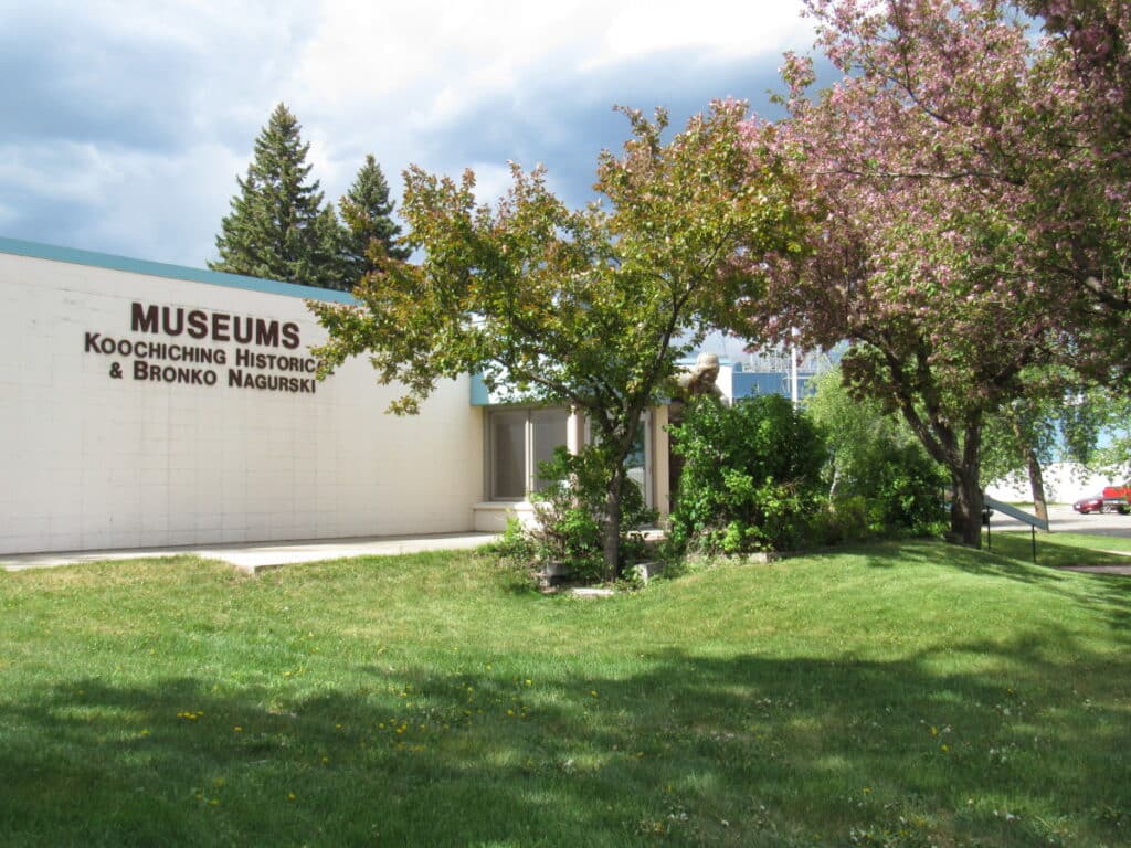 A building with a sign reading "Museums Koochiching Historical & Bronko Nagurski" stands behind green grass and leafy trees near Rainy Lake in Minnesota, with flowering branches under a partly cloudy sky. | Rainy Lake, MN