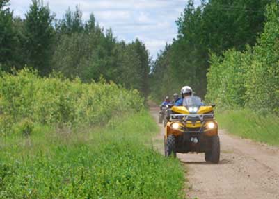 A person rides a yellow ATV on a dirt trail near Rainy Lake, surrounded by green trees and bushes, with two more riders following behind under a partly cloudy Minnesota sky. | Rainy Lake, MN