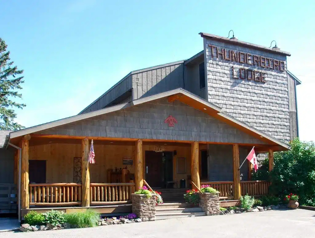 A rustic wooden lodge with a covered porch, flagpoles displaying American and Canadian flags, potted flowers, and a sign reading “Thunderbird Lodge” on the front. Surrounded by trees and shrubs, the building exudes the charm and spirit of Thunderbird tradition in a tranquil natural setting. | Rainy Lake, MN