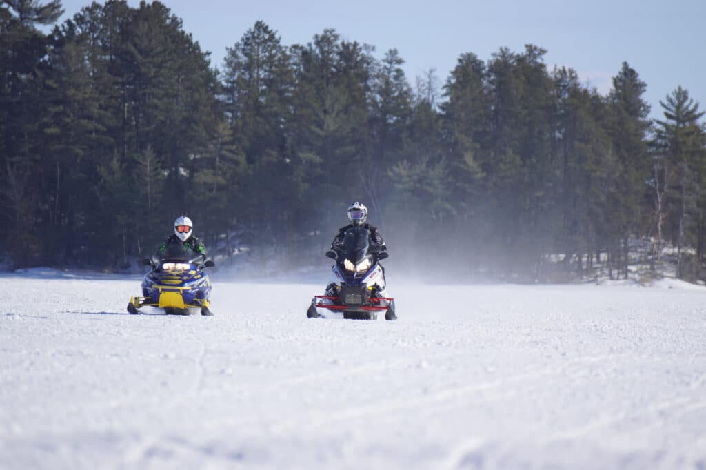 Two people riding snowmobiles across a snowy Minnesota landscape with trees in the background; both riders are wearing helmets and winter gear on a clear, sunny day. | Rainy Lake, MN