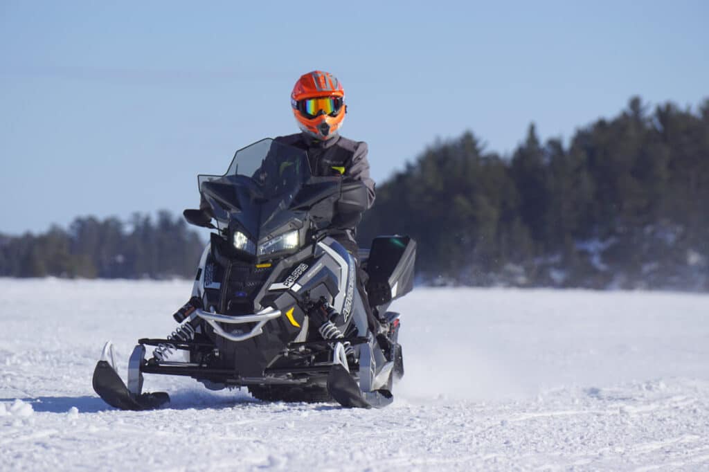 A person wearing an orange helmet and dark winter gear rides a black snowmobile across a snowy landscape near Rainy Lake, Minnesota, with trees in the background under a clear sky. | Rainy Lake, MN