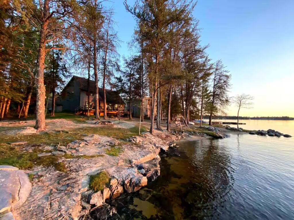 A cozy cabin surrounded by tall trees sits on a rocky shoreline at Rainy Lake, Minnesota, next to a calm lake at sunset, with golden sunlight illuminating the scene. | Rainy Lake, MN