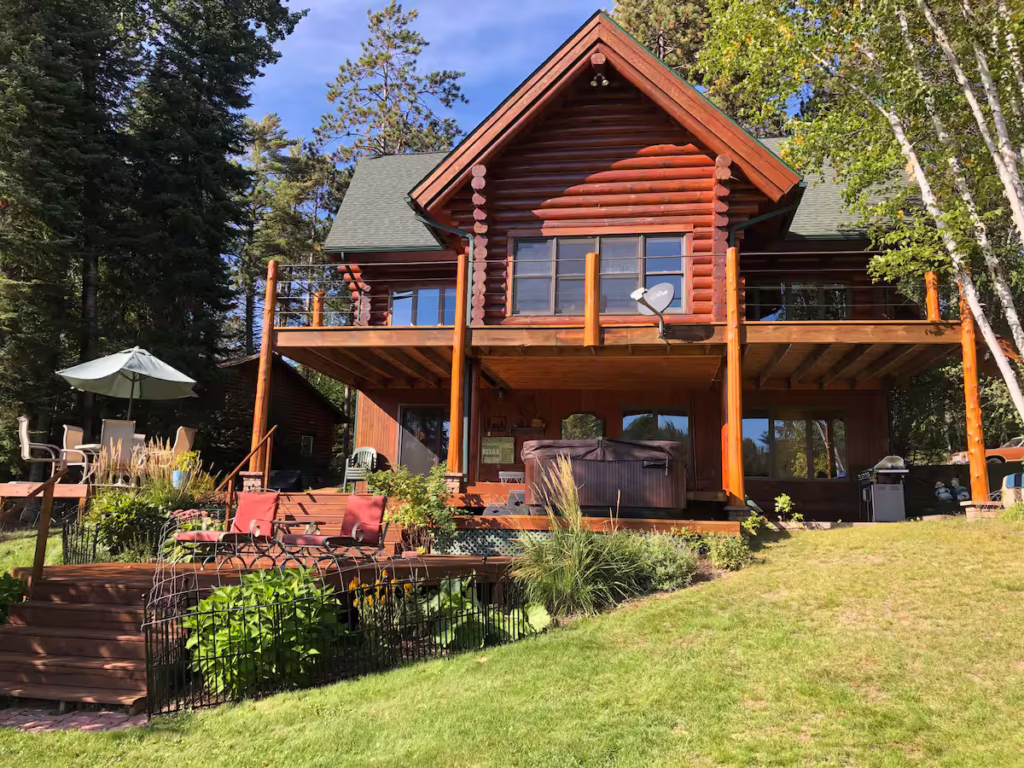 A log cabin at Twilight Bay with a large wooden deck, patio furniture, hot tub, and garden beds, surrounded by tall trees and green grass on a sunny day—your perfect AirBnB escape. | Rainy Lake, MN