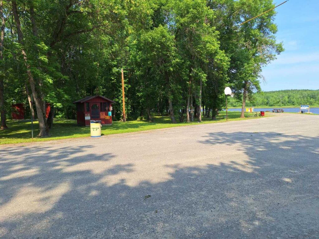 A small red cabin sits among trees near Rainy River and a tranquil lake. A basketball hoop, trash can, and picnic tables are visible on a paved area under a clear blue sky, making this spot in Nelson Park perfect for outdoor gatherings. | Rainy Lake, MN