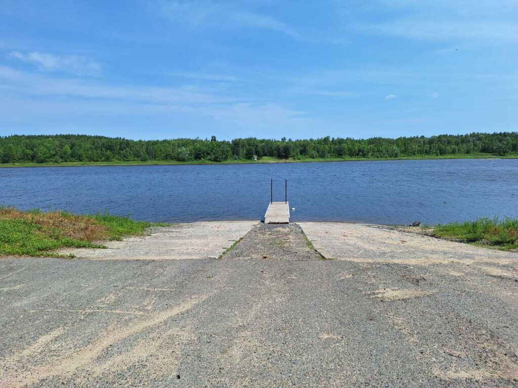A small dock extends from a concrete ramp into the calm waters of Rainy River, surrounded by green trees under a clear blue sky in Nelson Park. | Rainy Lake, MN