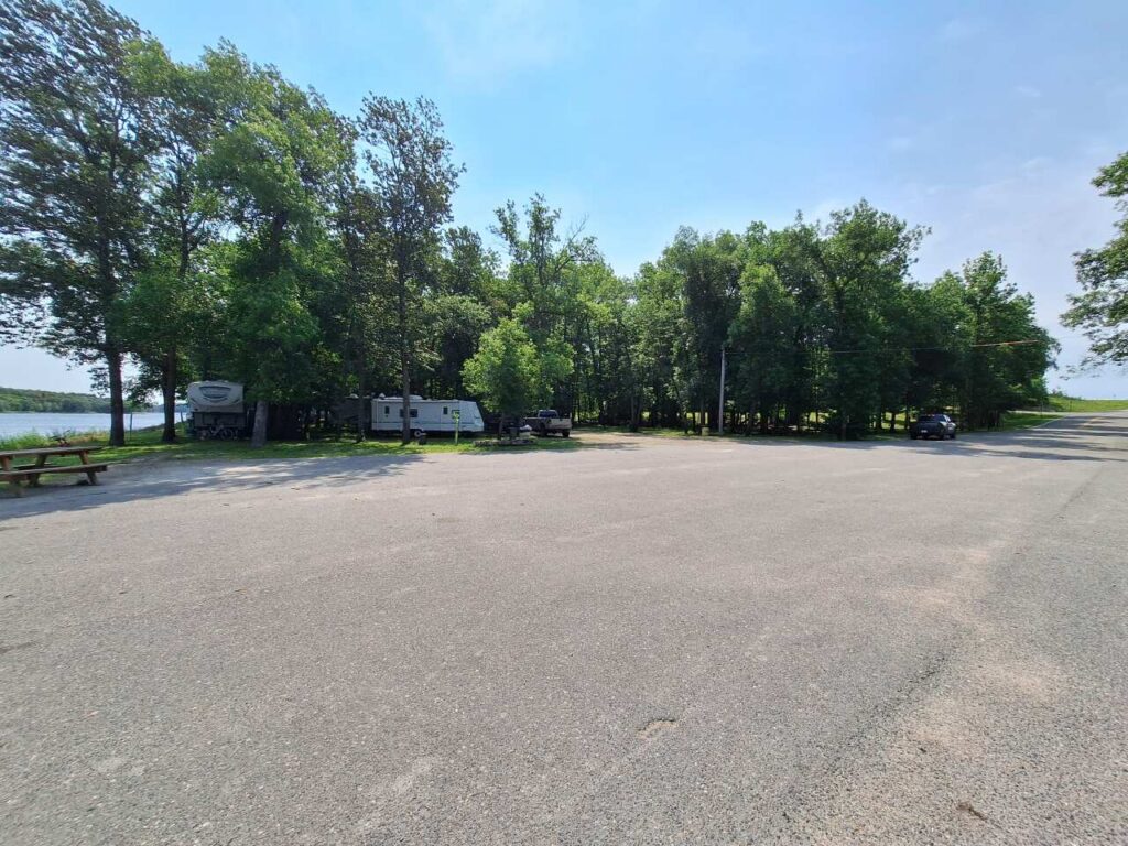 A mostly empty parking lot with a few vehicles and campers parked near a tree line at Nelson Park. Trees surround the lot, and Rainy River is visible on the left side under a clear blue sky. | Rainy Lake, MN