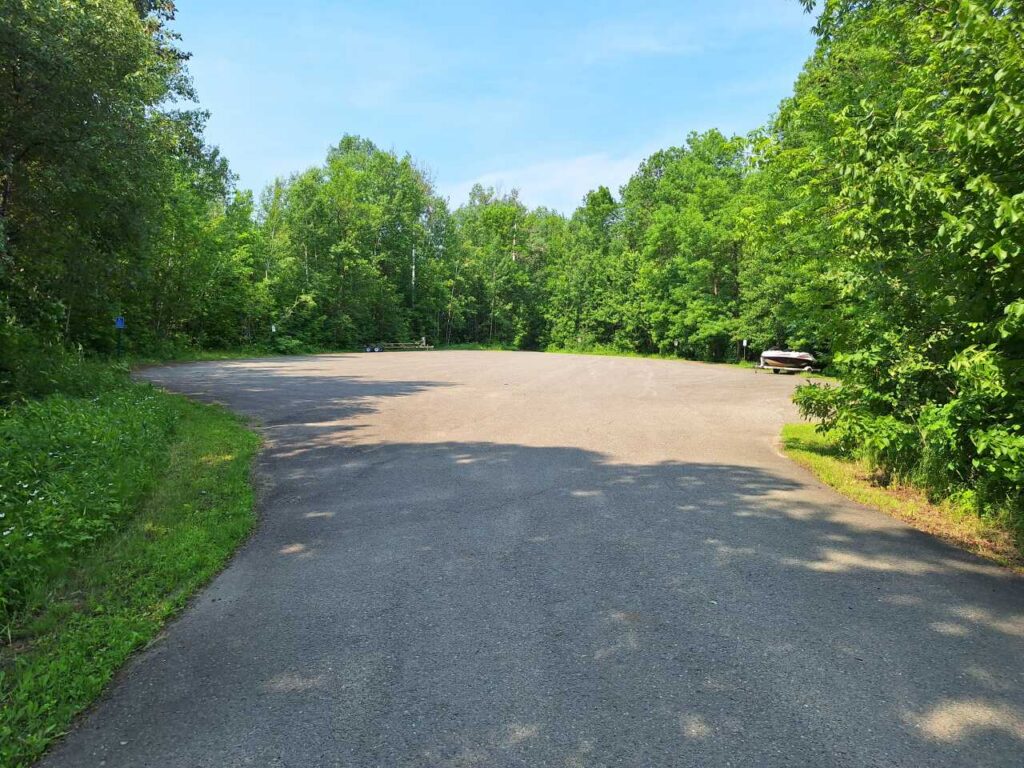 A mostly empty parking lot in Nelson Park, surrounded by trees under a clear blue sky, with a single parked vehicle visible on the right side and the tranquil waters of Rainy River nearby. | Rainy Lake, MN