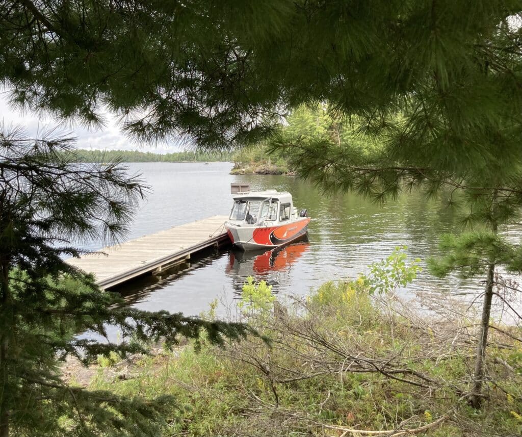 A small red and gray boat is tied to a wooden dock on Rainy Lake, its calm waters in Minnesota framed by green pine branches and foliage in the foreground. Trees and a cloudy sky are visible in the background. | Rainy Lake, MN