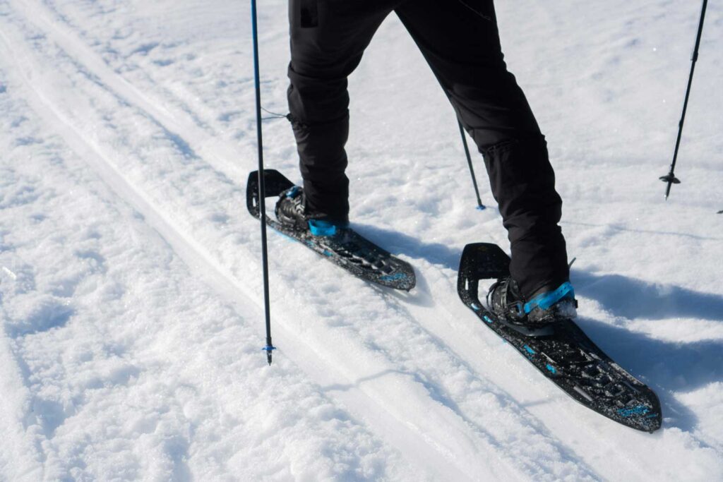 Person wearing black snowshoes and black pants walking on packed snow near Rainy Lake, Minnesota, using poles for support. The snow shows tracks from previous travelers. | Rainy Lake, MN