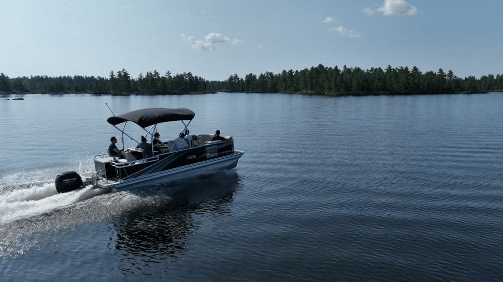 A group of people ride a pontoon boat with a canopy across calm Rainy Lake in Minnesota, leaving a gentle wake behind. Trees and a clear blue sky are visible in the background. | Rainy Lake, MN