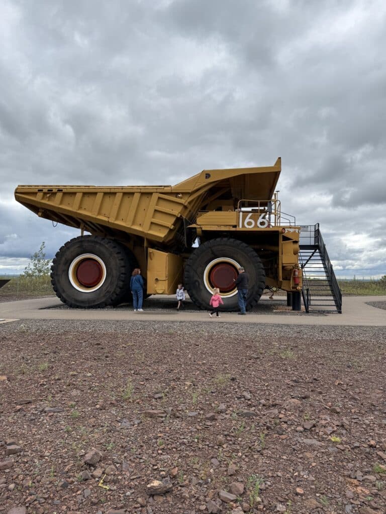 A large yellow mining dump truck is parked outdoors on gravel near Rainy Lake, Minnesota. Four people, including two children, stand by the truck, highlighting its massive size beneath a cloudy sky. | Rainy Lake, MN
