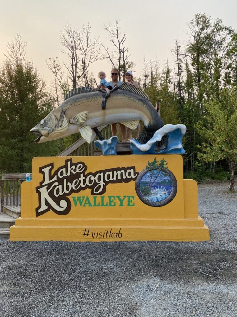 A family sits on top of a large walleye fish statue at the "Lake Kabetogama Walleye" sign in Minnesota, surrounded by trees, with "#visitkab" written at the bottom. | Rainy Lake, MN