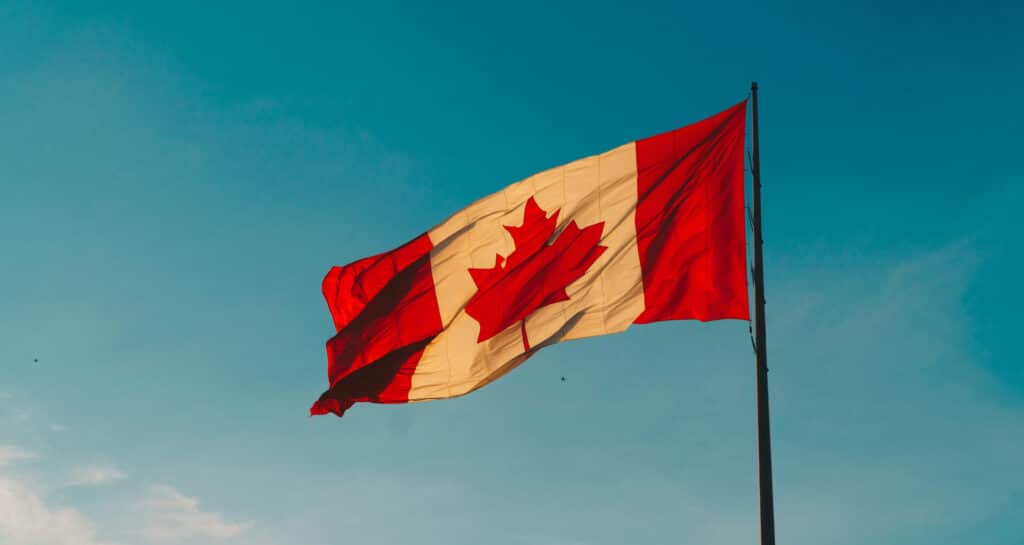 A Canadian flag with a red maple leaf in the center, flanked by red vertical bars on both sides, waving on a flagpole against a blue sky near Rainy Lake, just north of Minnesota. | Rainy Lake, MN