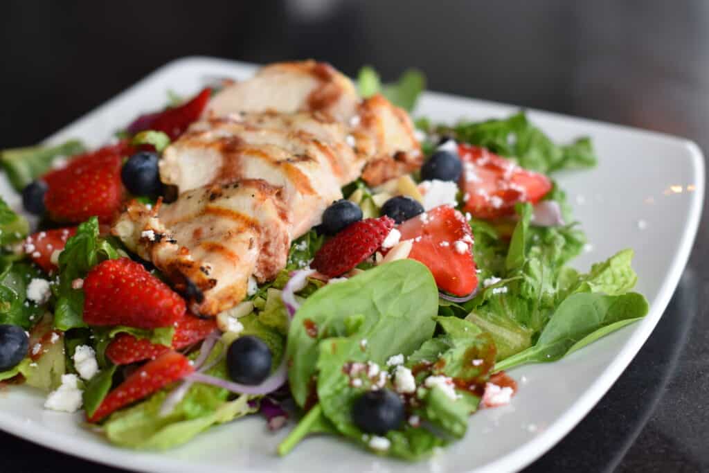 A fresh salad with leafy greens, grilled chicken breast, sliced strawberries, blueberries, red onions, crumbled feta cheese, and a drizzle of dressing—bringing a taste of Minnesota’s Rainy Lake to your table—served on a white square plate. | Rainy Lake, MN