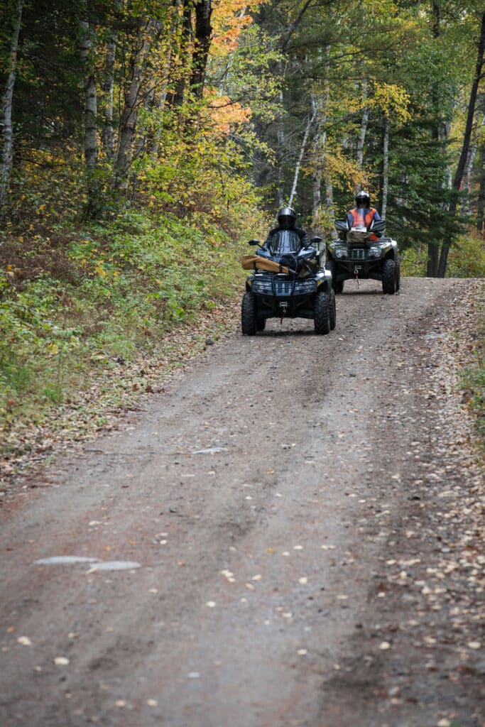 Two people ride ATVs up a dirt path in a Minnesota forest, surrounded by autumn foliage and trees with green and yellow leaves. | Rainy Lake, MN