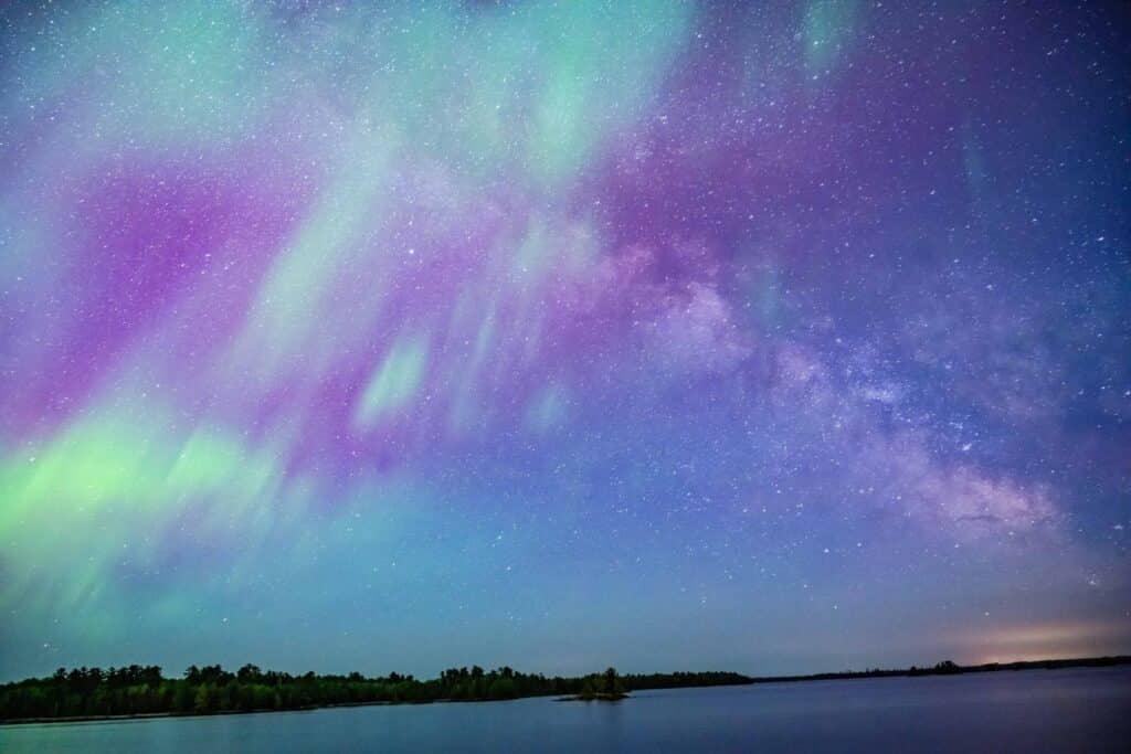 The northern lights with streaks of green, purple, and blue illuminate a star-filled night sky above Minnesota’s tranquil Rainy Lake and a dark, tree-lined horizon. | Rainy Lake, MN