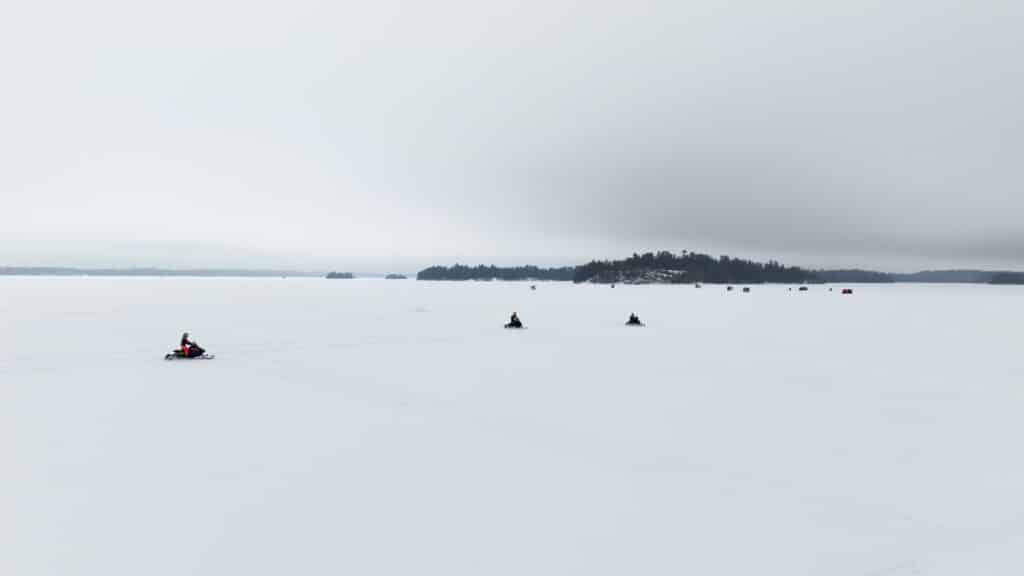 Three people ride snowmobiles across a vast snowy landscape on Minnesota's Rainy Lake, passing small dark huts with a distant tree-covered island beneath the cloudy sky. | Rainy Lake, MN