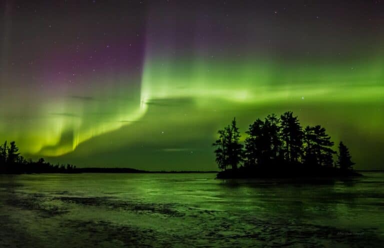 Green and purple northern lights illuminate the night sky above Rainy Lake, Minnesota, with a silhouette of a tree-covered island in the foreground. The water reflects the vibrant aurora colors, creating a serene, magical scene. | Rainy Lake, MN